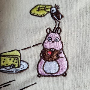 May include: Embroidered design featuring a pink hamster holding a cupcake with a red cherry. A bird is perched above the hamster, holding a yellow object. A slice of cake sits on a plate. The design is on a beige fabric.