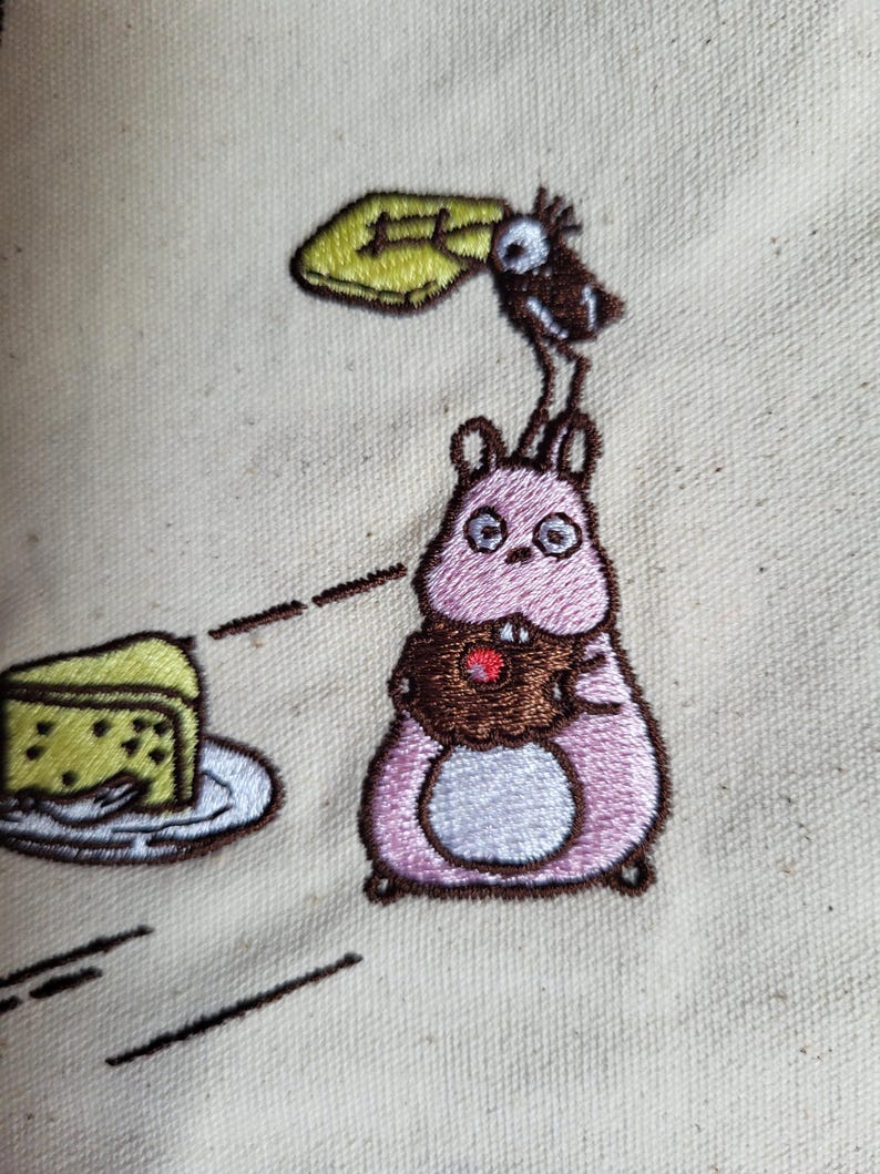 May include: Embroidered design featuring a pink hamster holding a cupcake with a red cherry. A bird is perched above the hamster, holding a yellow object. A slice of cake sits on a plate. The design is on a beige fabric.