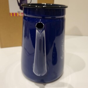 May include: A navy blue enamel coffee pot with a black rim and handle. The pot has a spout and a small opening near the top. The pot is sitting on a white surface. A cardboard box is in the background.