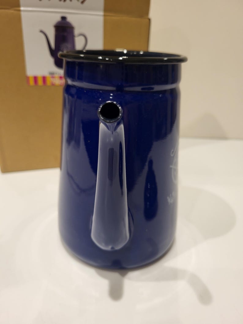May include: A navy blue enamel coffee pot with a black rim and handle. The pot has a spout and a small opening near the top. The pot is sitting on a white surface. A cardboard box is in the background.