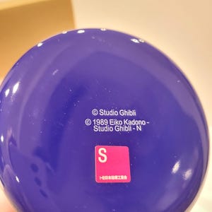May include: A close-up of a deep blue, spherical object with white text that reads "Studio Ghibli" and "1989 Eiko Kadono - Studio Ghibli - N". A pink and white sticker with the letter "S" is also visible.
