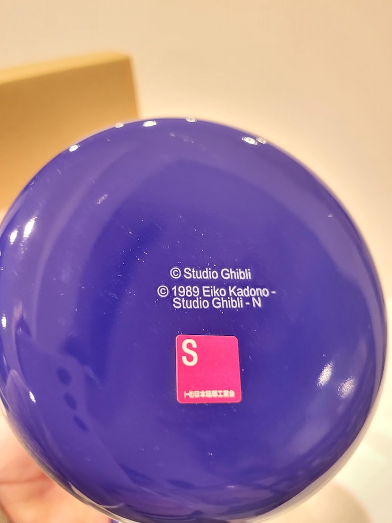 May include: A close-up of a deep blue, spherical object with white text that reads "Studio Ghibli" and "1989 Eiko Kadono - Studio Ghibli - N". A pink and white sticker with the letter "S" is also visible.