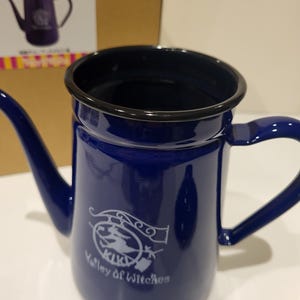 May include: A cobalt blue enamelware teapot with a black rim and handle. The teapot features a white graphic of a witch on a broom and the text "Valley of Witches". The teapot is displayed in front of its packaging.