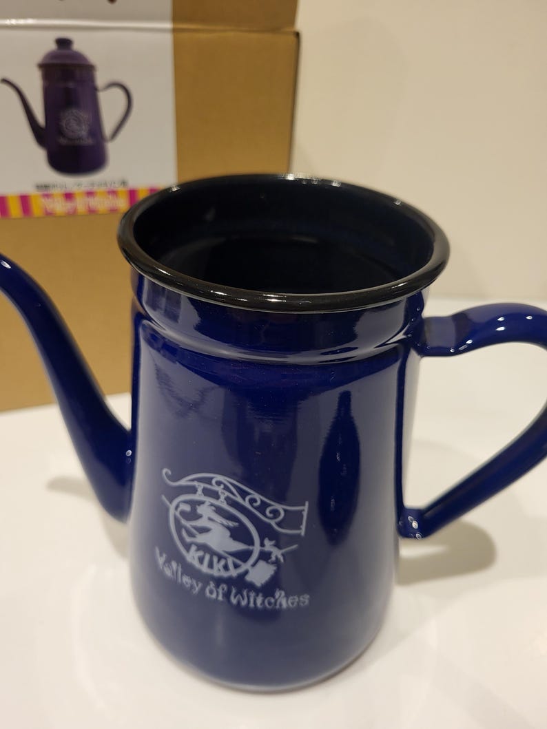 May include: A cobalt blue enamelware teapot with a black rim and handle. The teapot features a white graphic of a witch on a broom and the text "Valley of Witches". The teapot is displayed in front of its packaging.