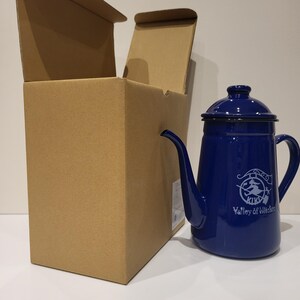 May include: A cobalt blue enamelware coffee pot with a black rim and handle. The pot has a long spout and a lid. The words "KIKI Valley of Witches" are printed on the side in white. The pot is next to an open cardboard box.