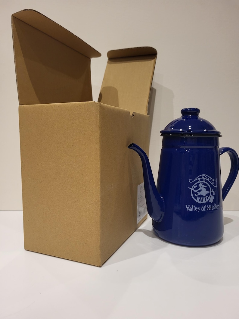 May include: A cobalt blue enamelware coffee pot with a black rim and handle. The pot has a long spout and a lid. The words "KIKI Valley of Witches" are printed on the side in white. The pot is next to an open cardboard box.