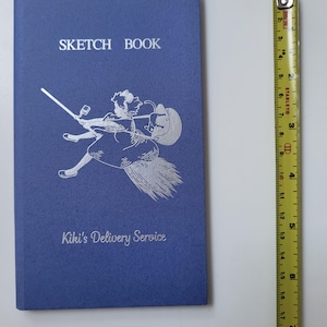 May include: A blue sketchbook with the words "SKETCH BOOK" at the top and "Kiki's Delivery Service" at the bottom. A silver illustration of a girl on a broom is in the center. A yellow and black measuring tape is to the right.