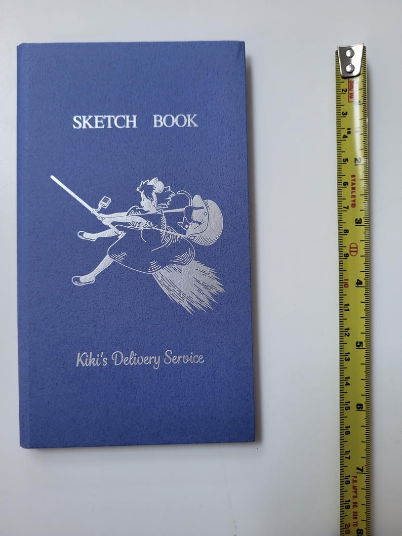 May include: A blue sketchbook with the words "SKETCH BOOK" at the top and "Kiki's Delivery Service" at the bottom. A silver illustration of a girl on a broom is in the center. A yellow and black measuring tape is to the right.
