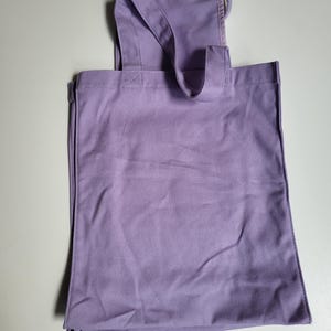 May include: A lavender tote bag with a rectangular body and a handle that extends over the top. The bag has a simple design and appears to be made of a sturdy fabric. A small tag is attached to the handle.