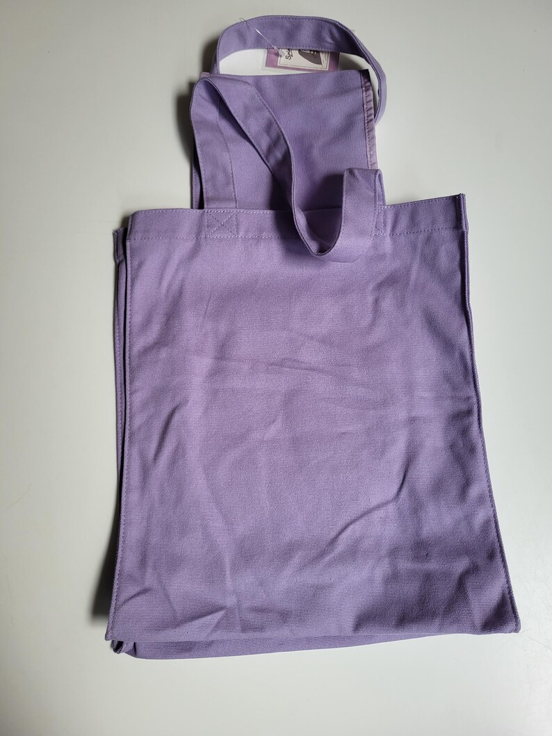 May include: A lavender tote bag with a rectangular body and a handle that extends over the top. The bag has a simple design and appears to be made of a sturdy fabric. A small tag is attached to the handle.