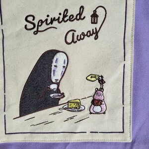 May include: A beige square patch with the words "Spirited Away" in brown script. The patch features an embroidered image of a character with a white mask and a black figure, a pink creature, and a slice of cake.