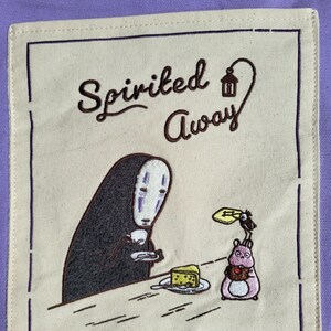 May include: A beige square patch with a dark purple border features an embroidered scene from "Spirited Away." The design includes the text "Spirited Away," a character with a white mask, and a pink creature holding a chocolate.