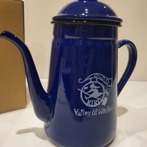May include: A cobalt blue enamel coffee pot with a long spout and a handle. The pot has a lid and a white graphic of a witch on a broomstick with the text "KIKI Valley of Witches".