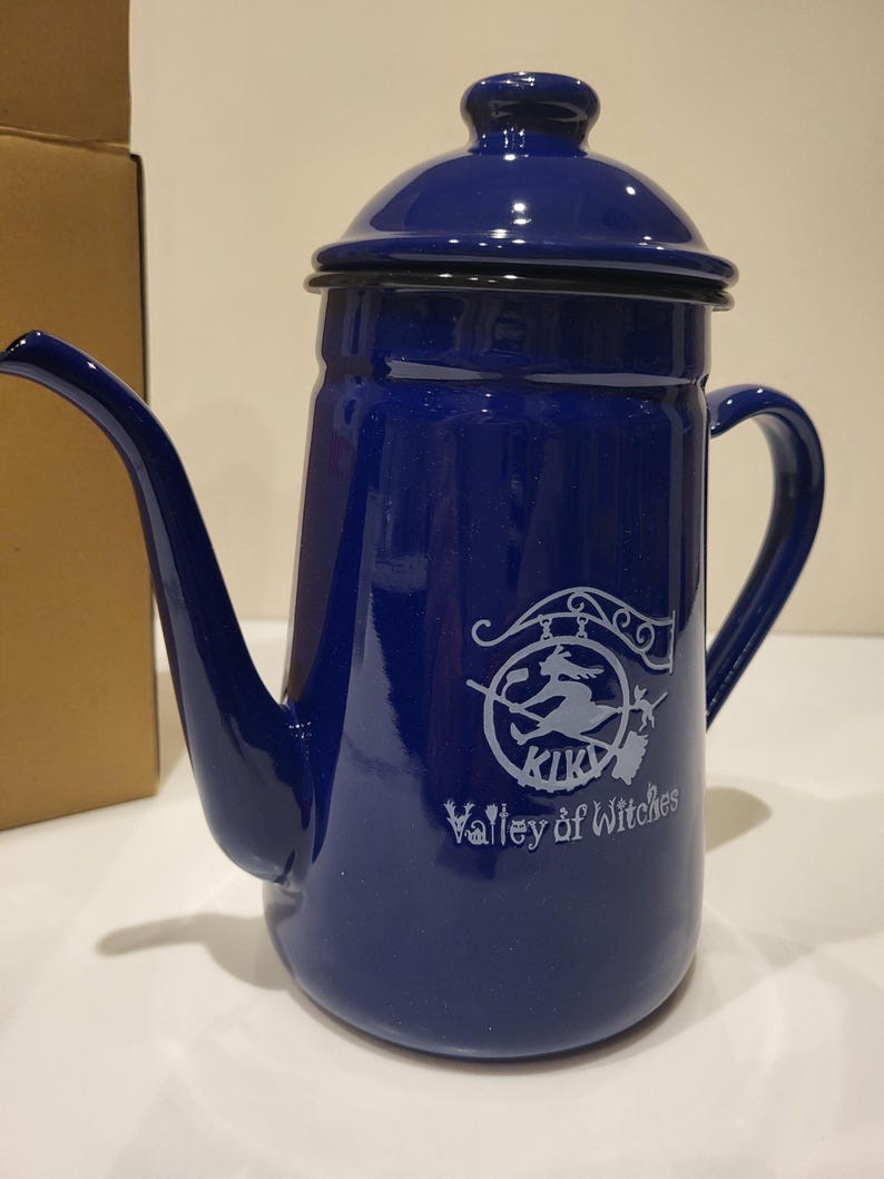 May include: A cobalt blue enamel coffee pot with a long spout and a handle. The pot has a lid and a white graphic of a witch on a broomstick with the text "KIKI Valley of Witches".