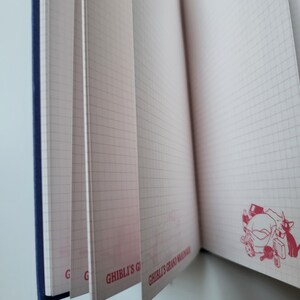 May include: Open notebook with grid-lined pages. The pages are pink and have a red stamp of a cartoon character. The notebook has a blue spine and the text "GHIBLI'S GRAND WAREHOUSE" is printed on the pages.