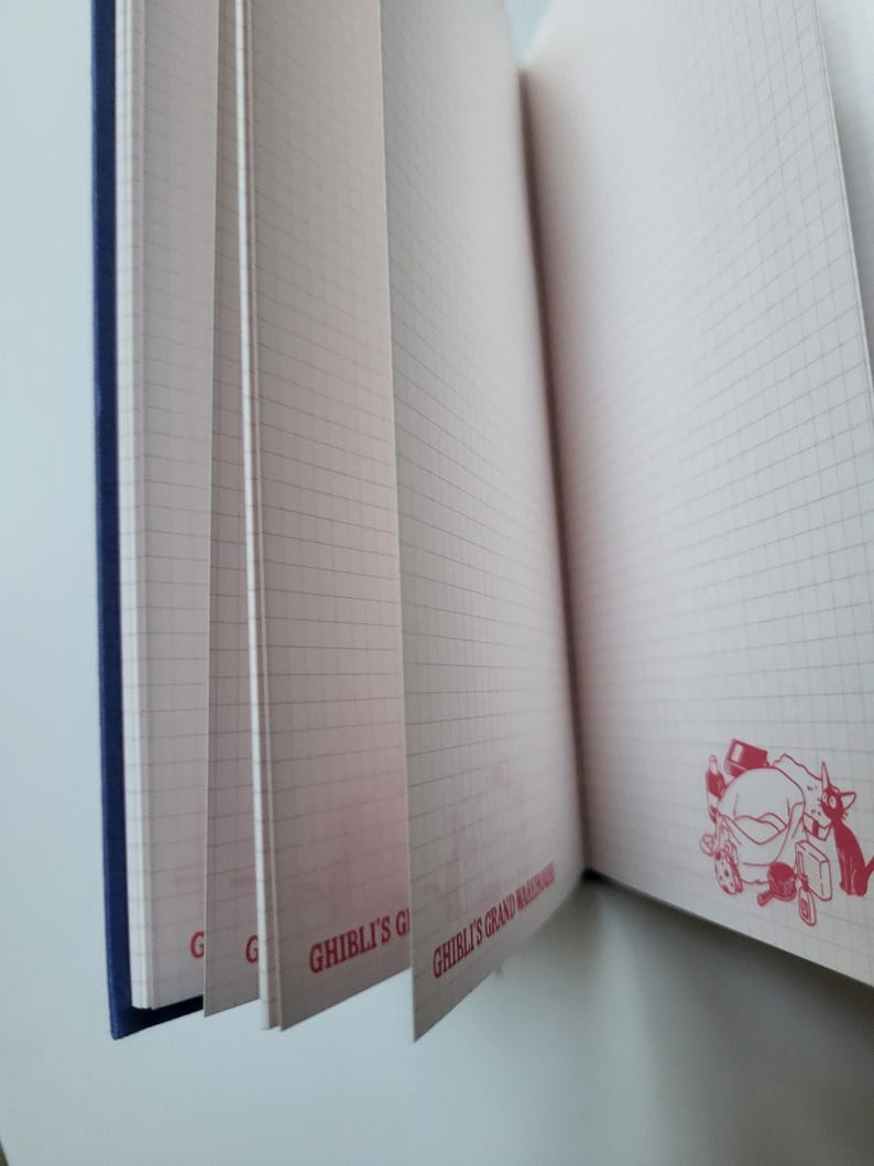 May include: Open notebook with grid-lined pages. The pages are pink and have a red stamp of a cartoon character. The notebook has a blue spine and the text "GHIBLI'S GRAND WAREHOUSE" is printed on the pages.