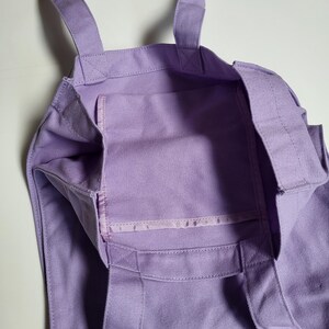 May include: A lavender tote bag with two handles and an interior pocket. The bag is made of a sturdy fabric and features reinforced stitching at the seams. The bag is designed for carrying items.