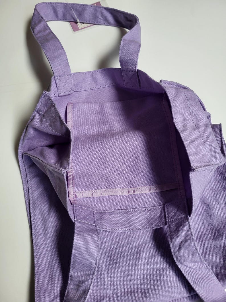May include: A lavender tote bag with two handles and an interior pocket. The bag is made of a sturdy fabric and features reinforced stitching at the seams. The bag is designed for carrying items.