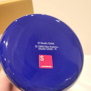 May include: A blue ceramic mug with a rounded base. The bottom of the mug features the text "&copy; Studio Ghibli" and a red sticker with the letter "S". The mug is likely a collectible item.