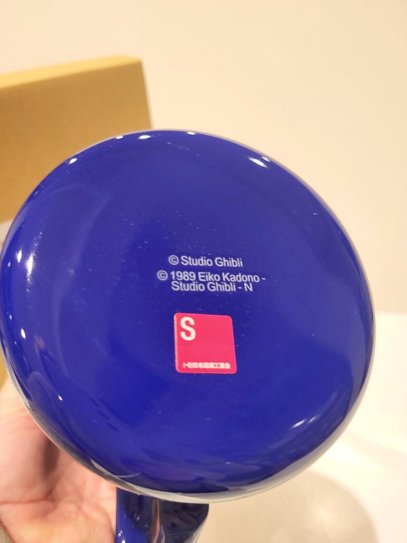 May include: A blue ceramic mug with a rounded base. The bottom of the mug features the text "&copy; Studio Ghibli" and a red sticker with the letter "S". The mug is likely a collectible item.