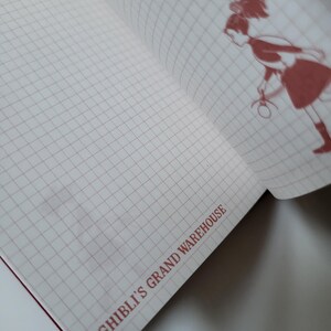 May include: Open notebook with a grid pattern and a red illustration of a character. The text "GHIBLI'S GRAND WAREHOUSE" is printed in red at the bottom of the page. The notebook has a red spine.