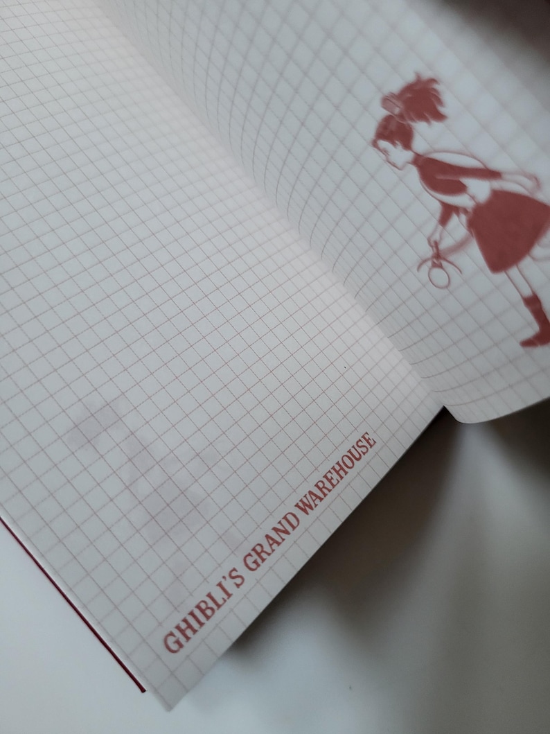May include: Open notebook with a grid pattern and a red illustration of a character. The text "GHIBLI'S GRAND WAREHOUSE" is printed in red at the bottom of the page. The notebook has a red spine.