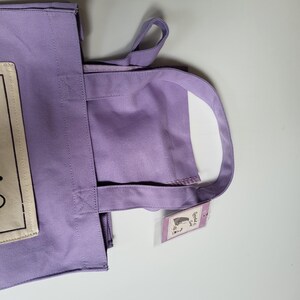May include: A lavender tote bag with matching handles. The bag has a rectangular patch with text on the front. A small tag with a graphic is attached to the handle. The bag is made of a sturdy fabric.