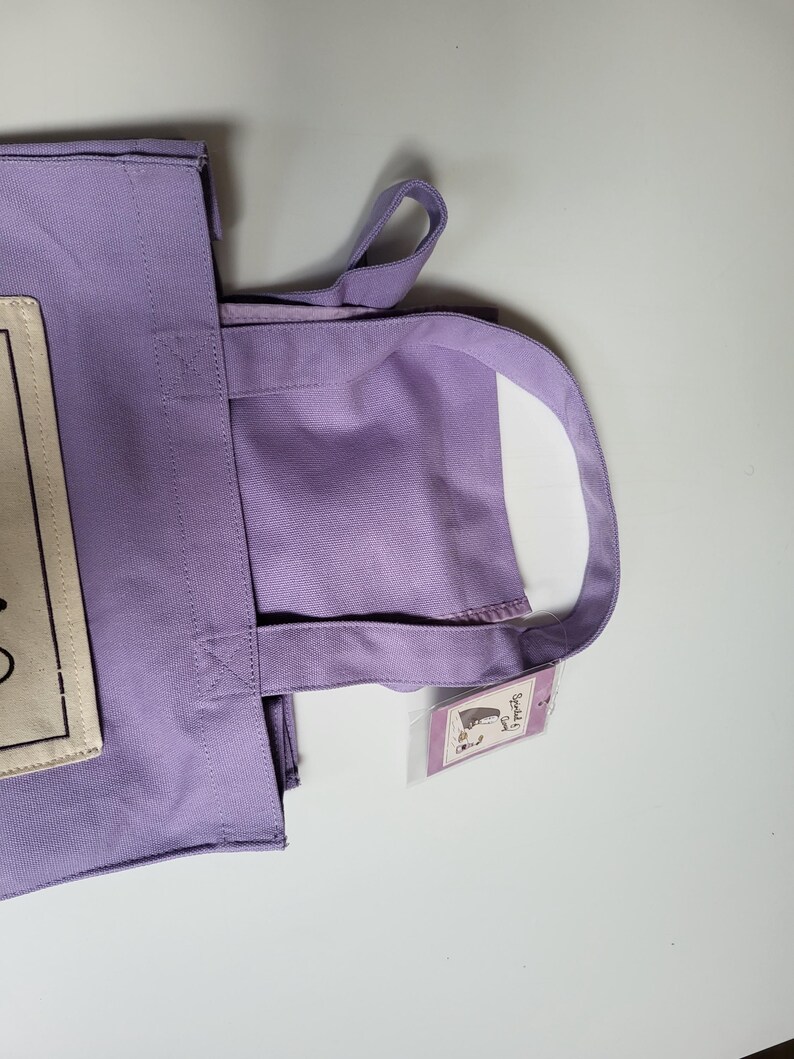 May include: A lavender tote bag with matching handles. The bag has a rectangular patch with text on the front. A small tag with a graphic is attached to the handle. The bag is made of a sturdy fabric.