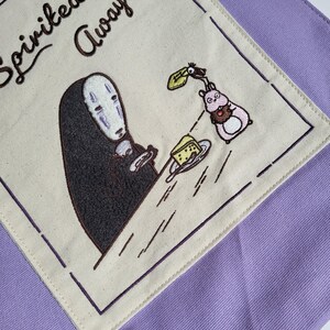 May include: A purple tote bag featuring an embroidered design from the movie "Spirited Away." The design includes the character No-Face holding a plate with a piece of cheese, and a pink creature with a chocolate treat. The words "Spirited Away" are embroidered in brown.