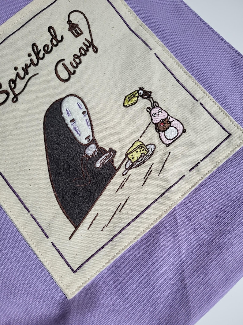 May include: A purple tote bag featuring an embroidered design from the movie "Spirited Away." The design includes the character No-Face holding a plate with a piece of cheese, and a pink creature with a chocolate treat. The words "Spirited Away" are embroidered in brown.