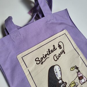 May include: A lavender tote bag with two handles. The bag features an embroidered patch with the words "Spirited Away" and an illustration of a character from the movie. The patch is cream-colored with a black border.