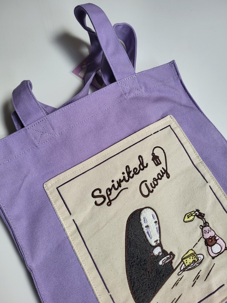 May include: A lavender tote bag with two handles. The bag features an embroidered patch with the words "Spirited Away" and an illustration of a character from the movie. The patch is cream-colored with a black border.