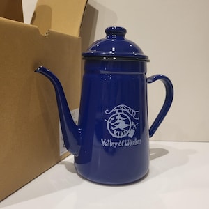 May include: A cobalt blue enamel coffee pot with a long spout, a curved handle, and a lid. The pot has a white graphic of a witch on a broom and the text "KIKI Valley of Witches". The pot is next to a cardboard box.