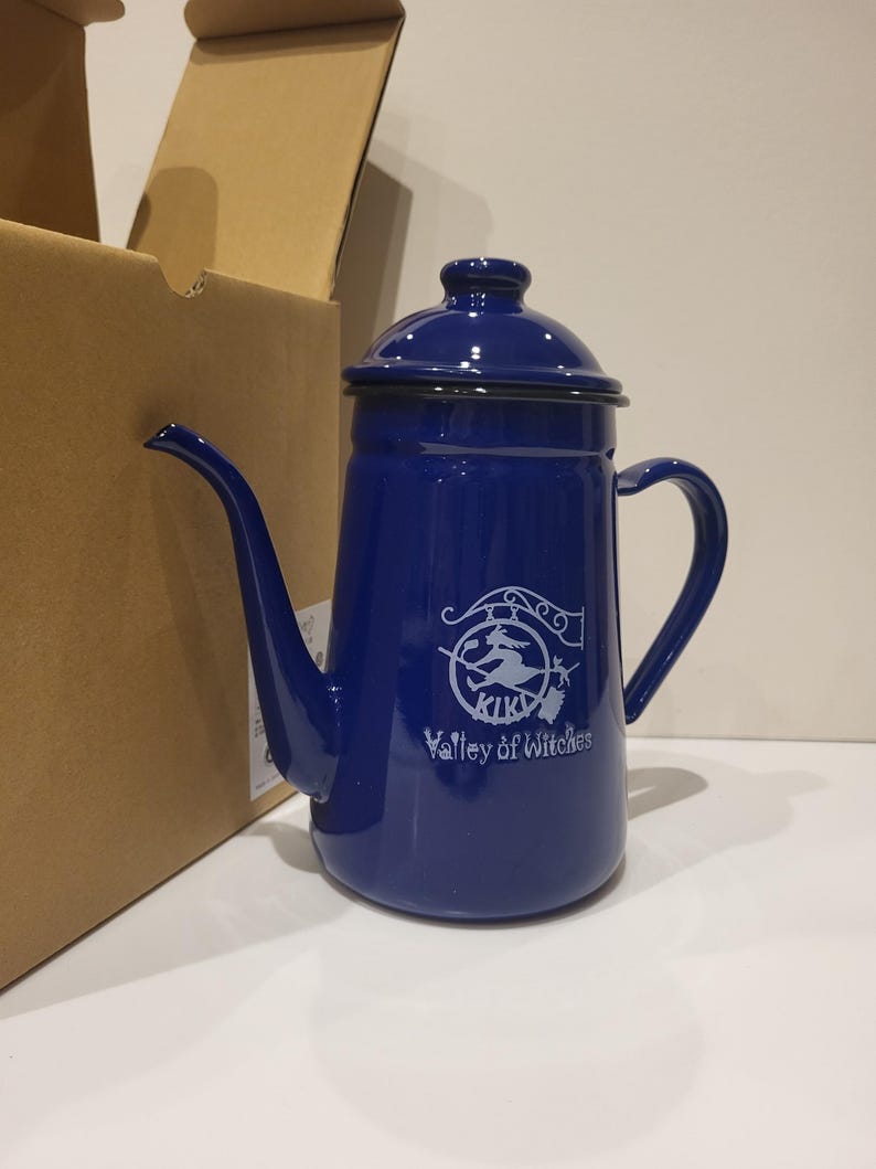 May include: A cobalt blue enamel coffee pot with a long spout, a curved handle, and a lid. The pot has a white graphic of a witch on a broom and the text "KIKI Valley of Witches". The pot is next to a cardboard box.