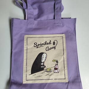 May include: A lavender tote bag featuring a graphic from the animated film "Spirited Away." The design includes the character No-Face and other elements from the movie. The bag has two handles and a rectangular design.