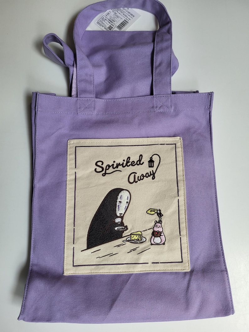May include: A lavender tote bag featuring a graphic from the animated film "Spirited Away." The design includes the character No-Face and other elements from the movie. The bag has two handles and a rectangular design.