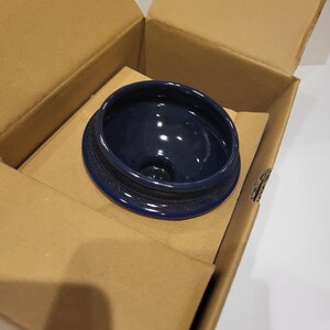 May include: A navy blue ceramic lid with a textured rim sits inside an open cardboard box. The lid has a glossy interior and a dark blue color. The box is made of corrugated cardboard and is open, revealing the lid.