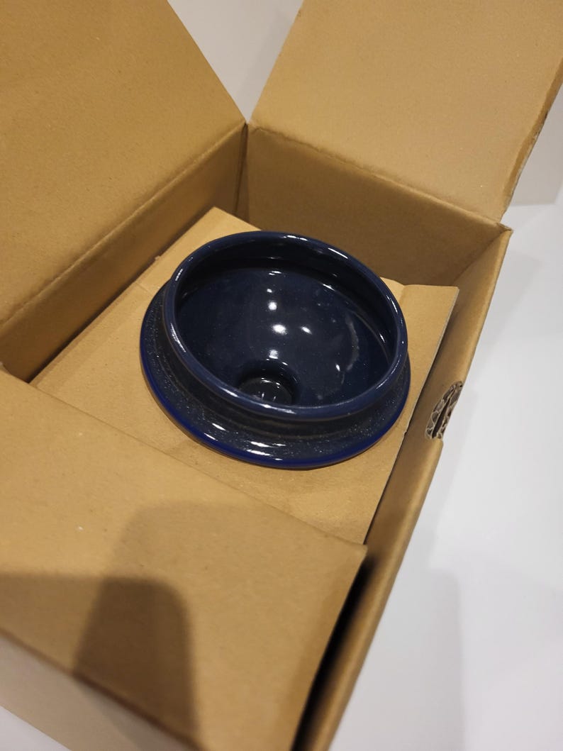 May include: A navy blue ceramic lid with a textured rim sits inside an open cardboard box. The lid has a glossy interior and a dark blue color. The box is made of corrugated cardboard and is open, revealing the lid.