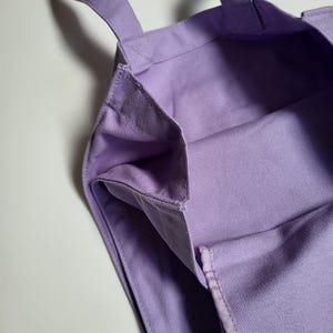 May include: A close-up of a lavender tote bag. The bag is made of a sturdy canvas material and features two handles. The interior of the bag is visible, showing the same lavender fabric. The bag is empty and sits on a white surface.