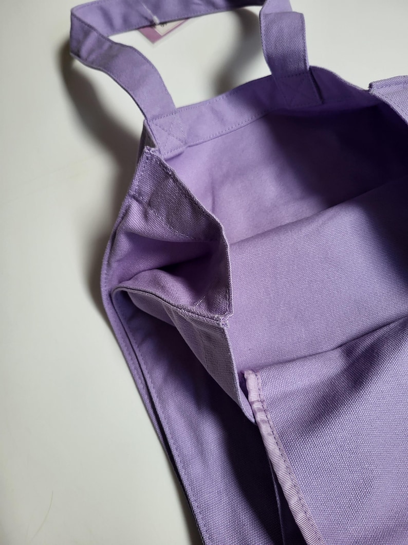 May include: A close-up of a lavender tote bag. The bag is made of a sturdy canvas material and features two handles. The interior of the bag is visible, showing the same lavender fabric. The bag is empty and sits on a white surface.