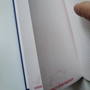May include: Open notebook with grid-lined pages. The pages are a light pink color with a grid pattern in a darker pink. The notebook has a blue spine. The text "GHIBLI'S GRAND WAREHOUSE" is printed in red on the bottom right corner of the page.