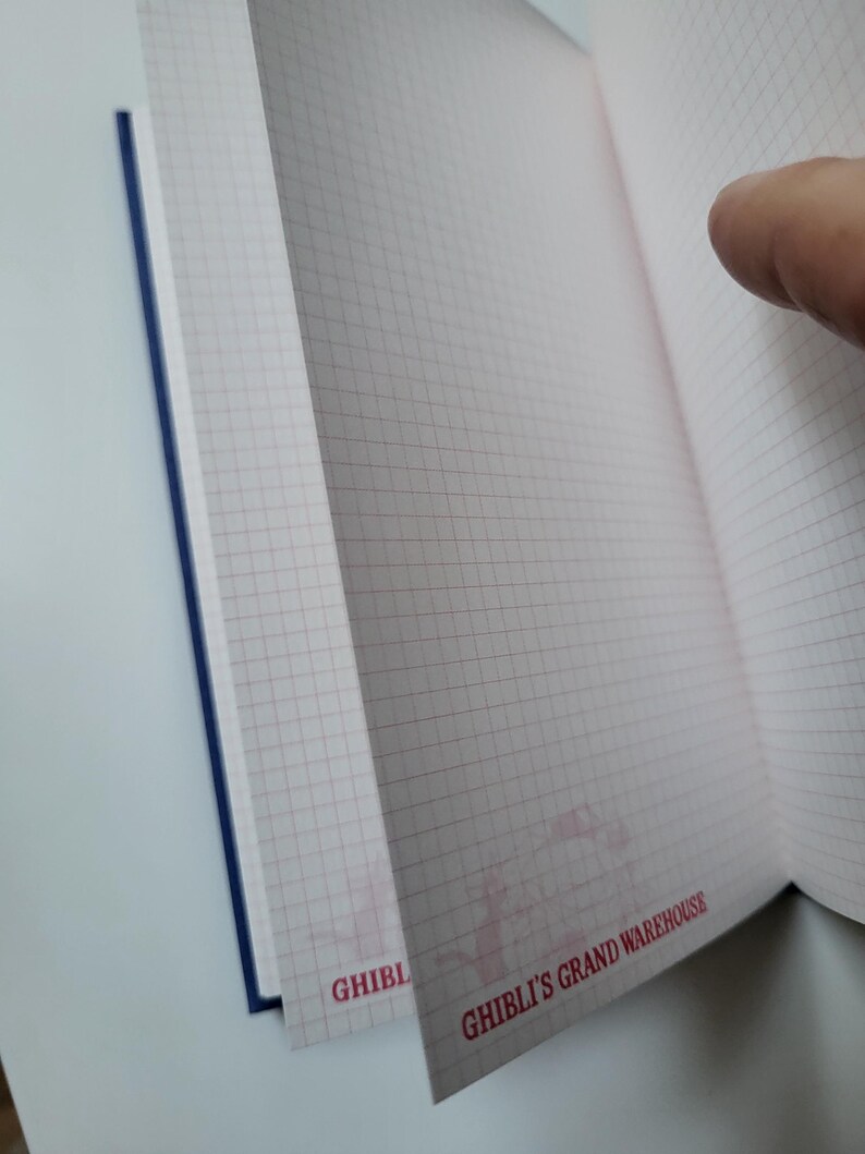 May include: Open notebook with grid-lined pages. The pages are a light pink color with a grid pattern in a darker pink. The notebook has a blue spine. The text "GHIBLI'S GRAND WAREHOUSE" is printed in red on the bottom right corner of the page.
