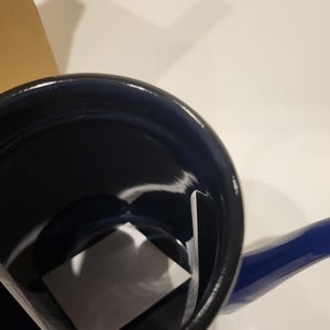 May include: A blue enamel saucepan with a matching handle. The interior is black, and a white square is inside the pan. The pan is sitting on a white surface.