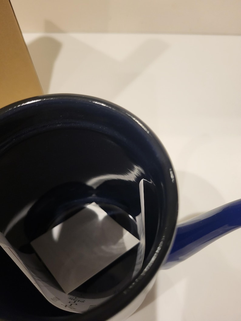 May include: A blue enamel saucepan with a matching handle. The interior is black, and a white square is inside the pan. The pan is sitting on a white surface.