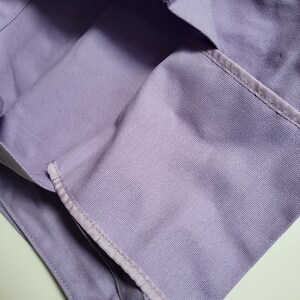May include: A close-up of a lavender tote bag made of a sturdy fabric. The bag has a rectangular shape with a reinforced top edge and a strap. The stitching is visible along the seams, and the bag appears to be empty.