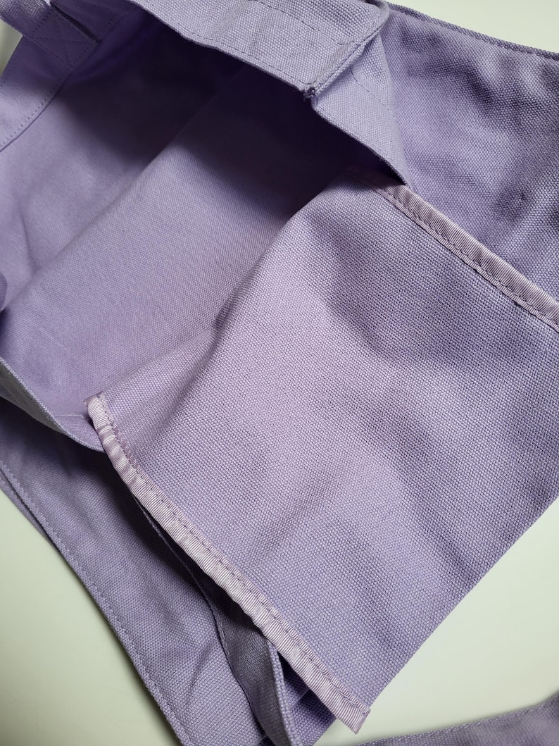 May include: A close-up of a lavender tote bag made of a sturdy fabric. The bag has a rectangular shape with a reinforced top edge and a strap. The stitching is visible along the seams, and the bag appears to be empty.