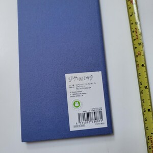 May include: A rectangular, textured, periwinkle-colored notebook with a white label featuring Japanese text and a green logo. A yellow measuring tape is positioned to the right of the notebook. The notebook is approximately 8 inches long.