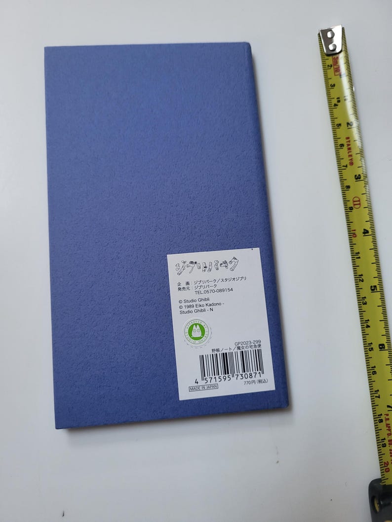 May include: A rectangular, textured, periwinkle-colored notebook with a white label featuring Japanese text and a green logo. A yellow measuring tape is positioned to the right of the notebook. The notebook is approximately 8 inches long.