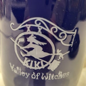 May include: A dark blue mug with a white graphic of a witch on a broomstick inside a circle, with the word "KIKI" above and "Valley of Witches" below. The mug has a glossy finish.