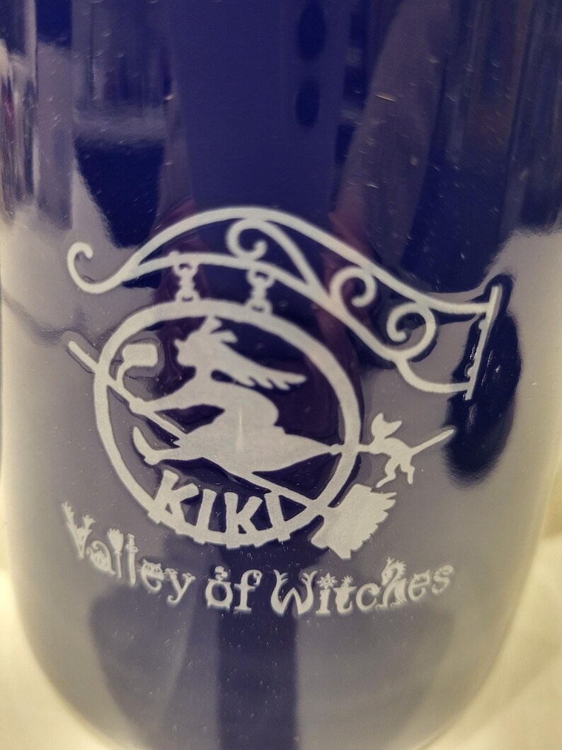 May include: A dark blue mug with a white graphic of a witch on a broomstick inside a circle, with the word "KIKI" above and "Valley of Witches" below. The mug has a glossy finish.